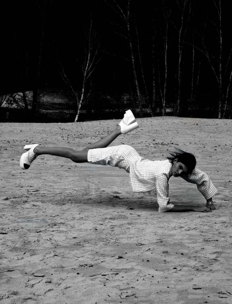 A person lying face down with legs raised and wearing high-heeled shoes and a patterned dress on a sandy surface outdoors.