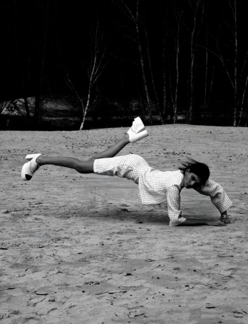 A person lying face down with legs raised and wearing high-heeled shoes and a patterned dress on a sandy surface outdoors.