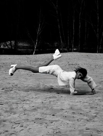 A person lying face down with legs raised and wearing high-heeled shoes and a patterned dress on a sandy surface outdoors.