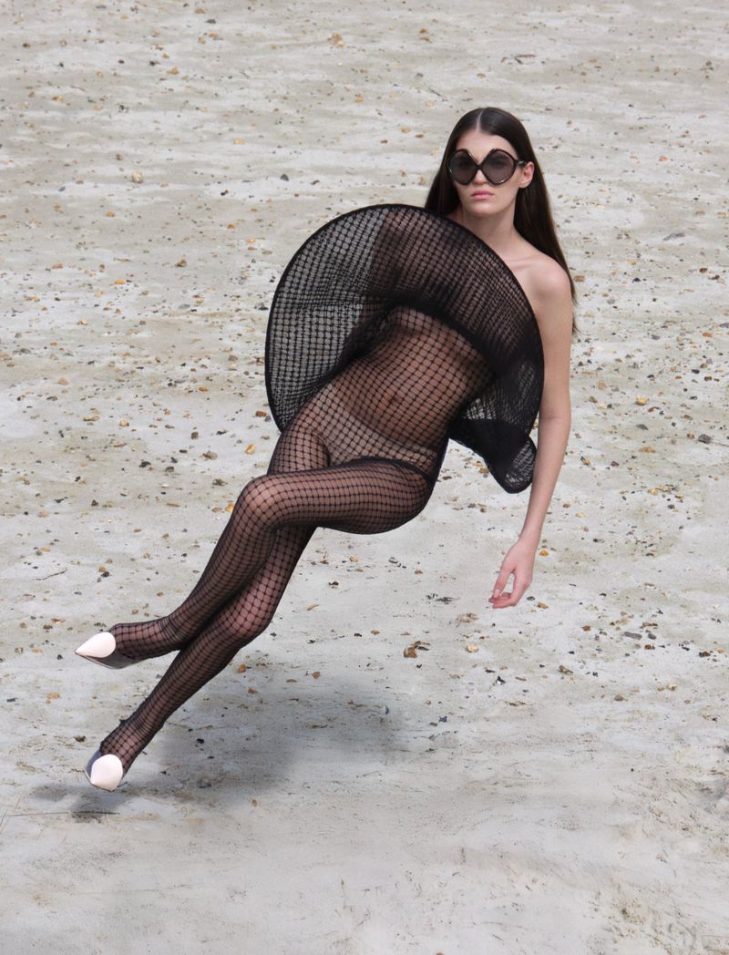 A woman in a black net bodysuit and geometric hat poses on a sandy surface, creating a fashion-forward and visually striking editorial image.