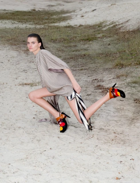 A woman poses on a sandy landscape wearing a draped dress with patterned heeled shoes. Neutral color palette fashion shot.