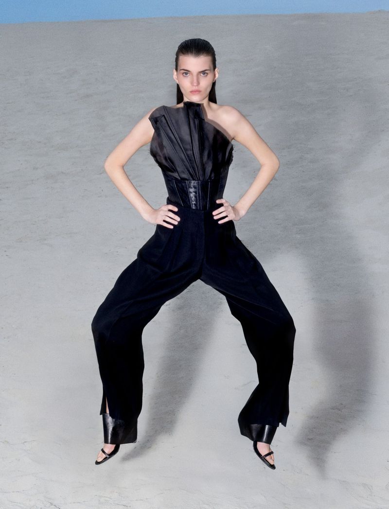 Fashion model in black one-shoulder jumpsuit striking confident pose in a stark studio environment against a light blue backdrop.