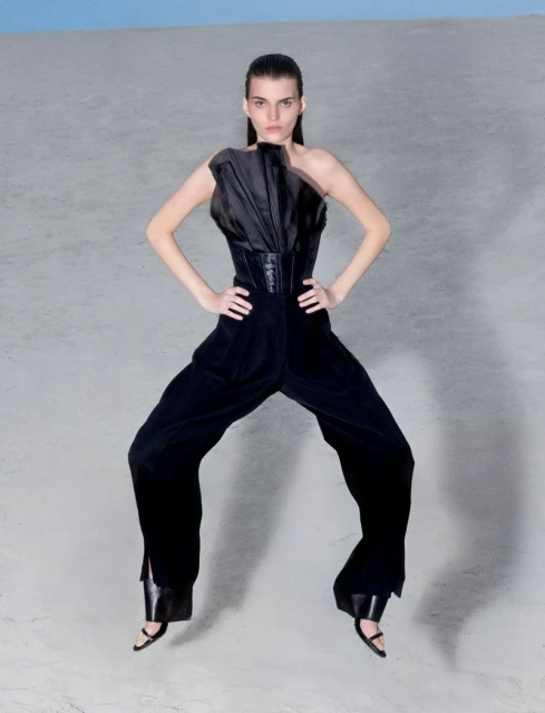 Fashion model in black one-shoulder jumpsuit striking confident pose in a stark studio environment against a light blue backdrop.