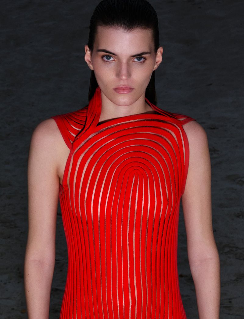 Woman in a red sleeveless dress poses in a studio. The dress features a unique, curved design. Shot at eye-level, the background is dark.