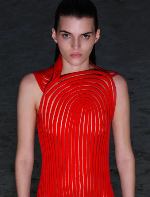 Woman in a red sleeveless dress poses in a studio. The dress features a unique, curved design. Shot at eye-level, the background is dark.