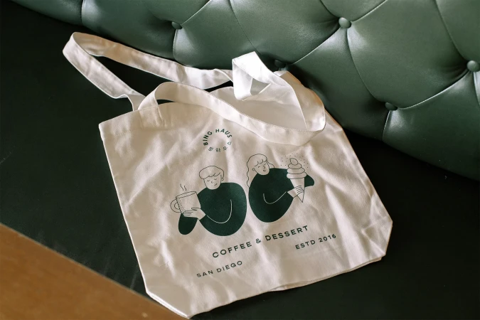 Cotton tote bag featuring minimalist drawings of people with coffee and ice cream on a tufted, dark-green surface.