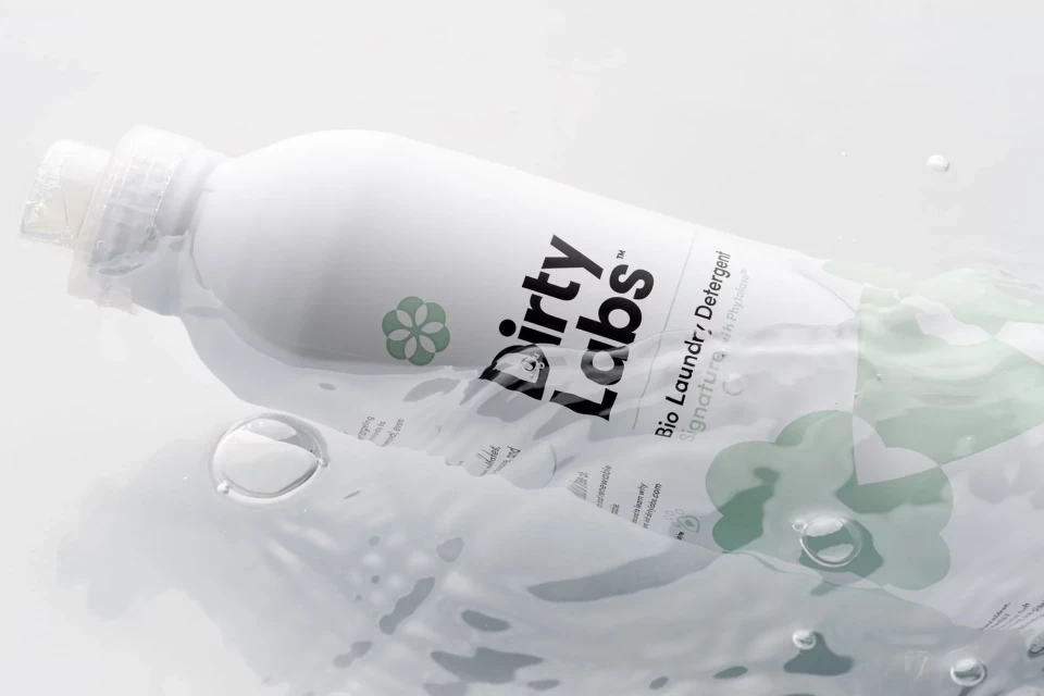 White bottle of Dirty Labs bio laundry detergent floats in clear water with bubbles, against a bright white studio background.