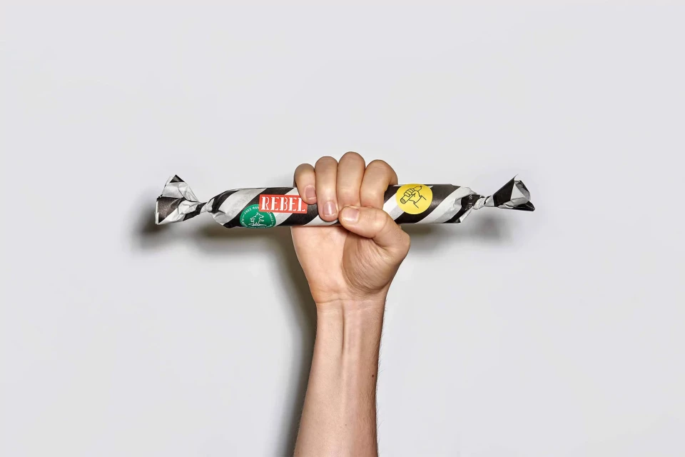 Arm holding a striped black and white wrapped candy, featuring the 'REBEL' logo, against a plain white background in a studio shot.