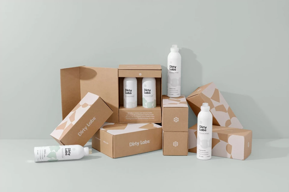 Several white Dirty Labs laundry detergent bottles and brown cardboard packaging boxes arranged on a light green surface in a studio setting.