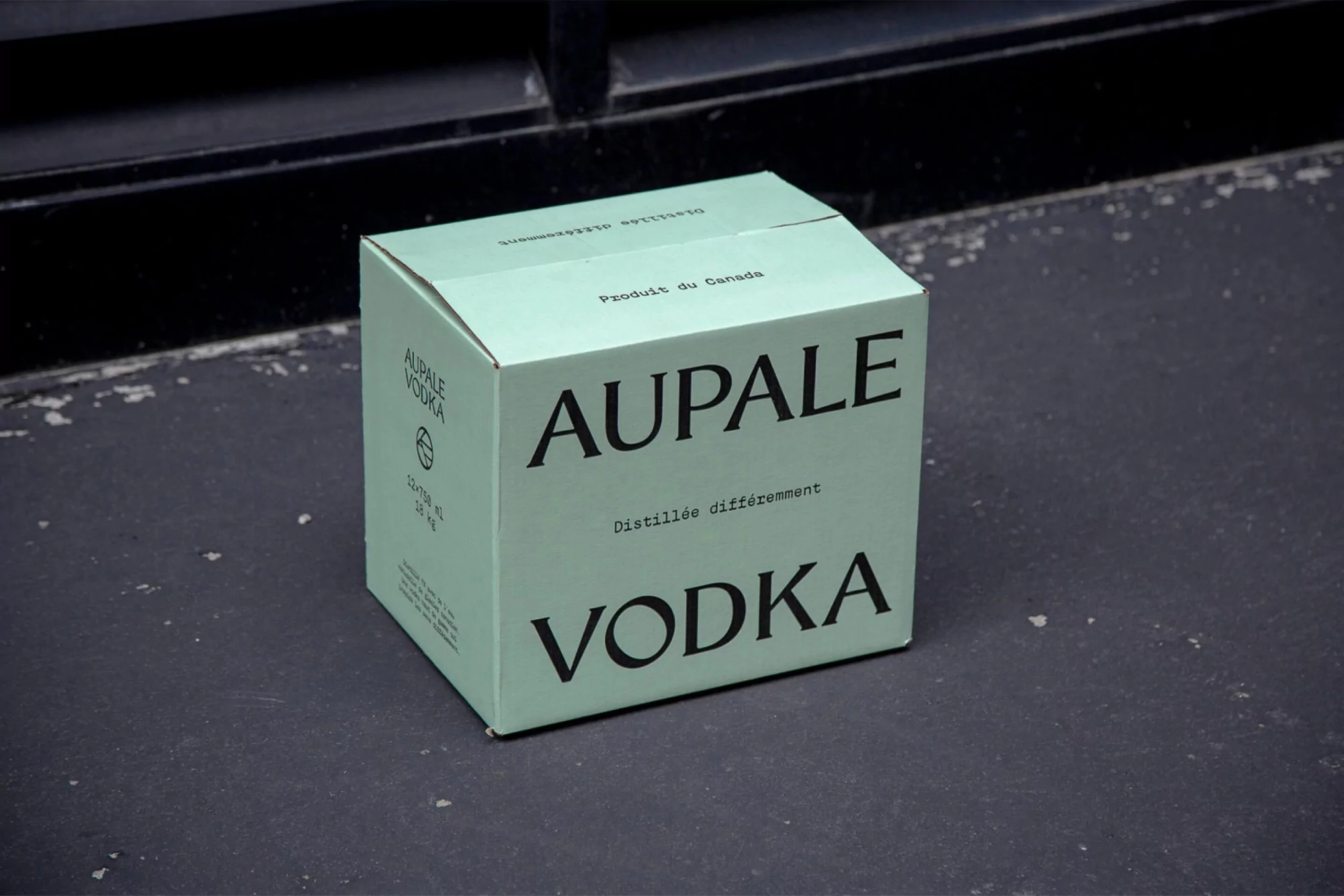 A light-green cardboard box with 'AUPALE VODKA' printed on it sits on a concrete surface against a dark background.