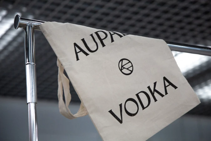 Canvas tote bag with 'AUPARA VODKA' printed in black on a metal clothing rack against a blurred grey background.