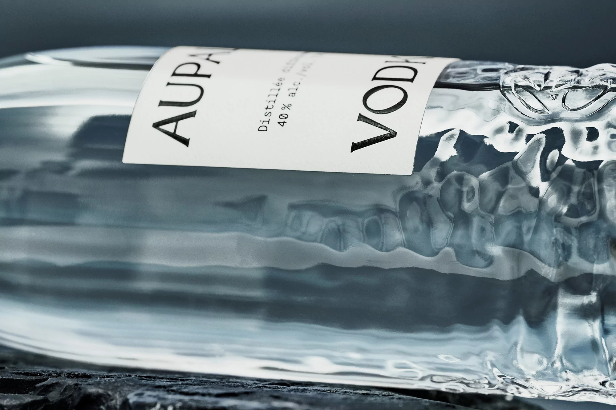 Close-up of a clear, textured glass bottle with a white label reading 'AUPALE VODKA' against a blurred dark backdrop.