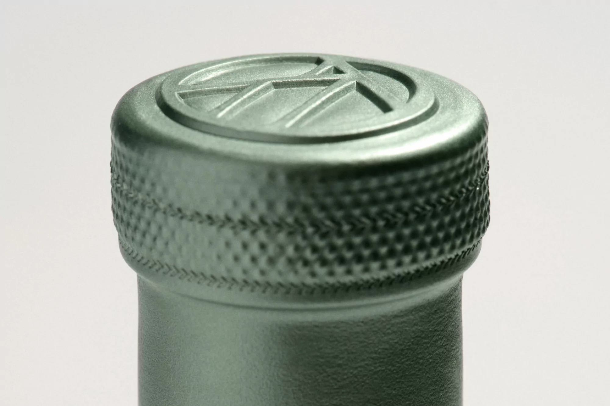 Close-up shot of a bottle neck and cap with a geometric logo, detailed texture, and a muted green color against a white background.