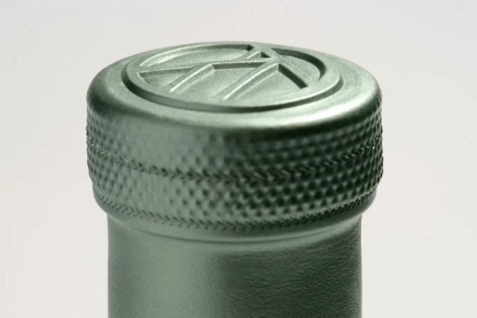 Close-up shot of a bottle neck and cap with a geometric logo, detailed texture, and a muted green color against a white background.