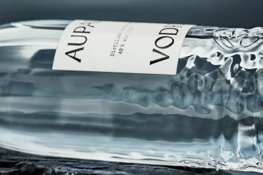 Close-up of a clear glass vodka bottle with a white label reading AUPALE VODKA resting on a dark textured surface.