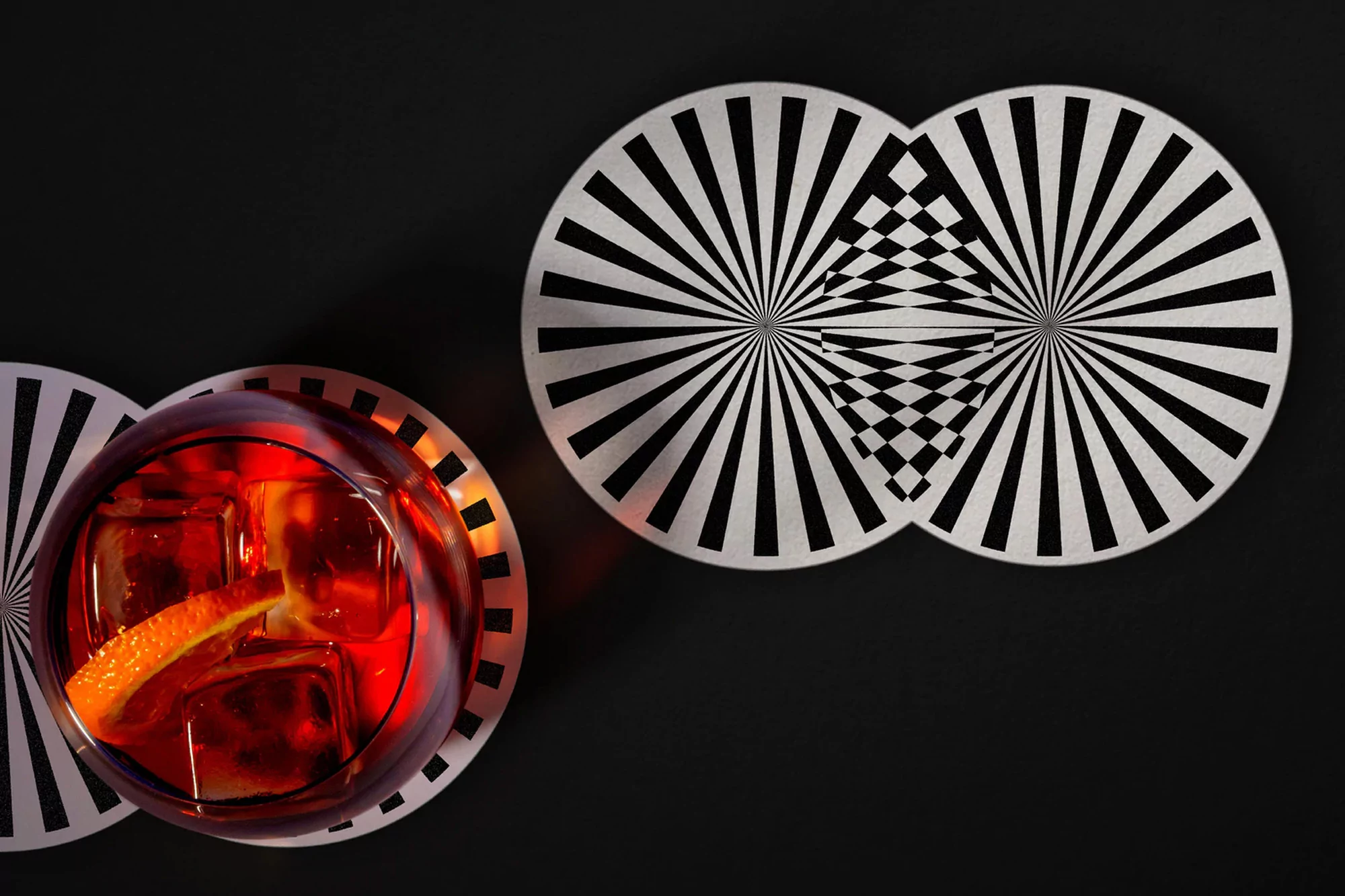 Overhead shot of a red cocktail with ice and orange peel on a round coaster with a black background, geometric pattern, and studio lighting.