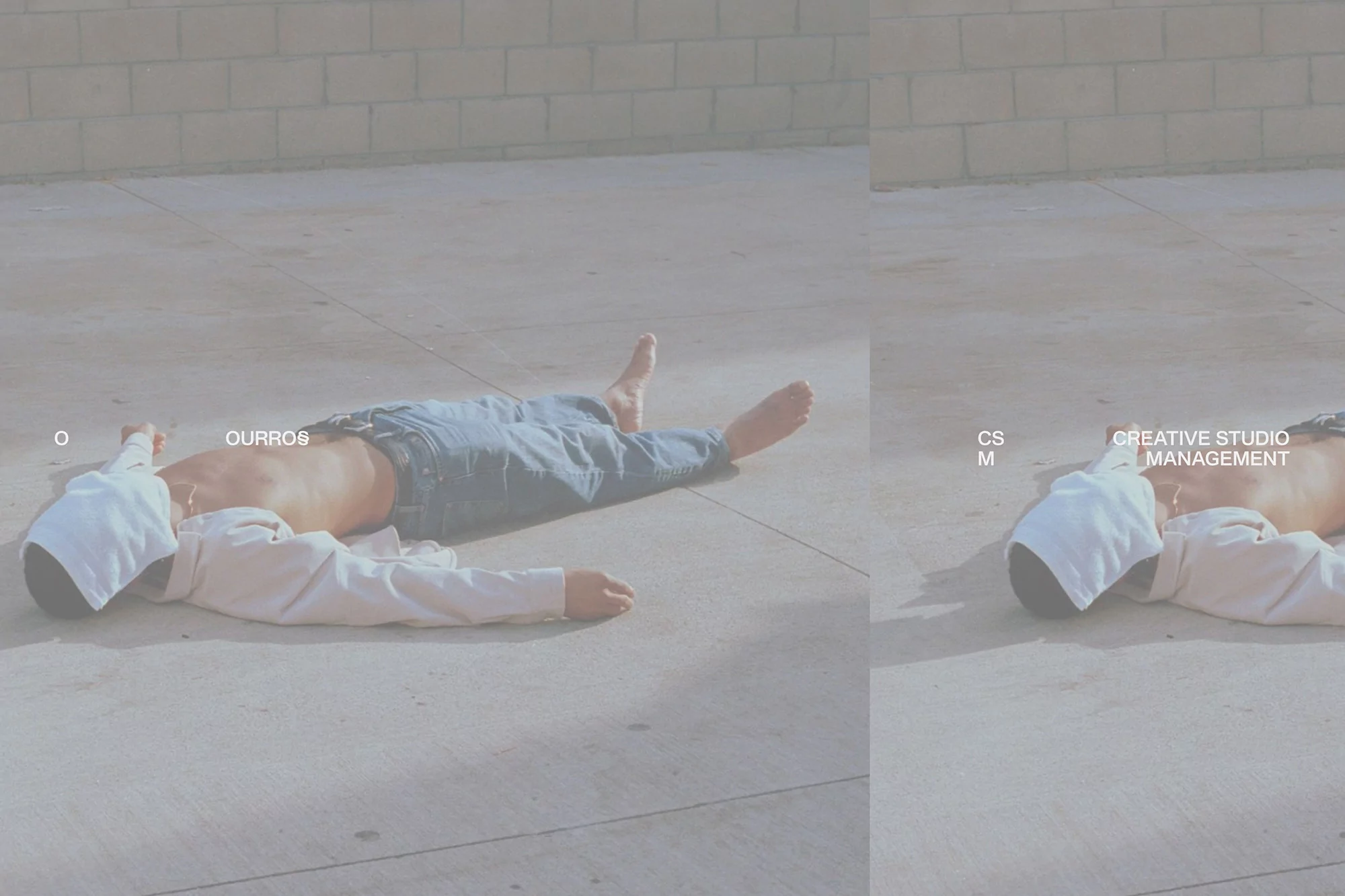Barefoot person lies on pavement in jeans and long-sleeve top with a white hood against a cinderblock wall in full sunlight.