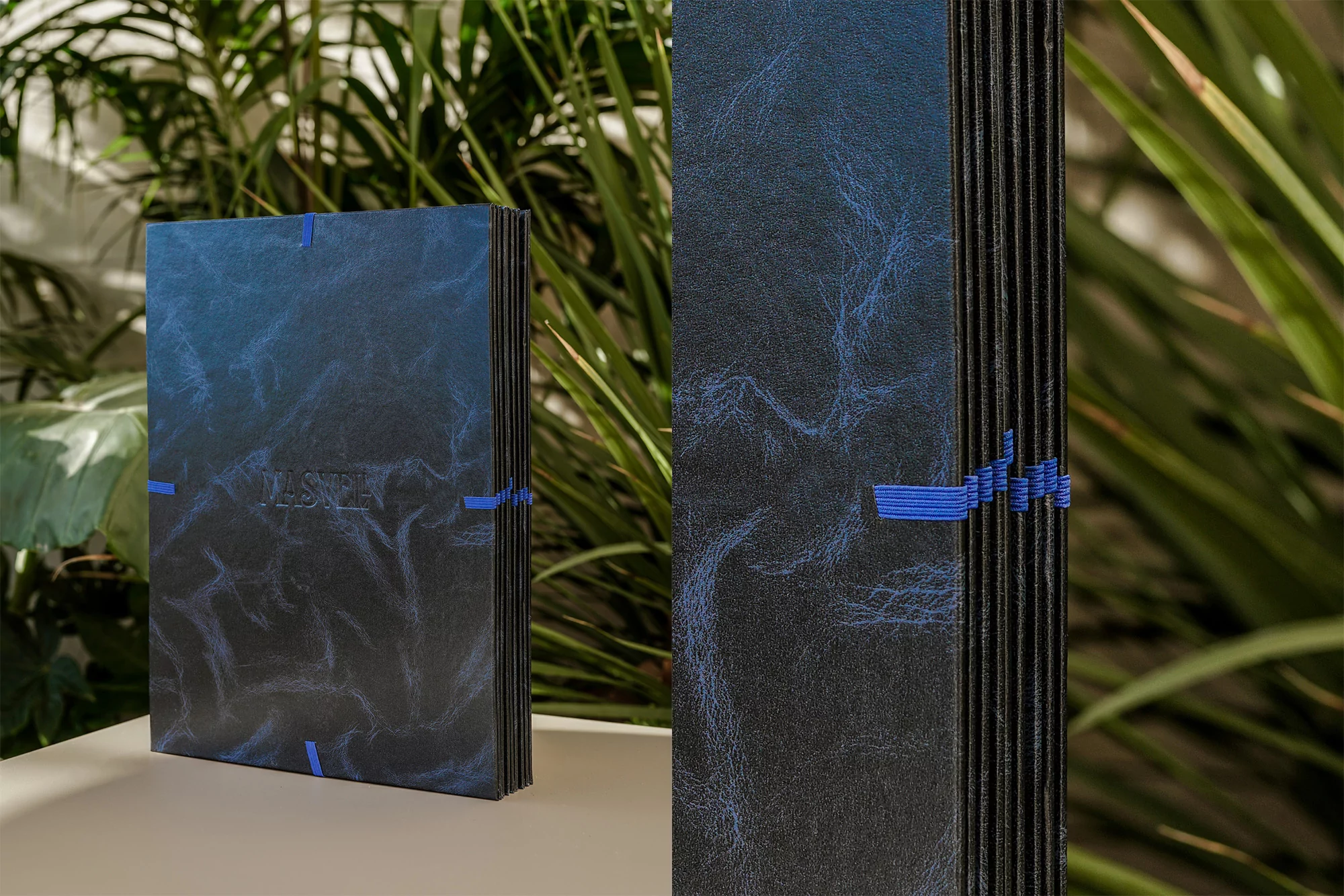 Stack of dark blue books with textured covers and blue bands, set against a soft background of green plants in natural light.