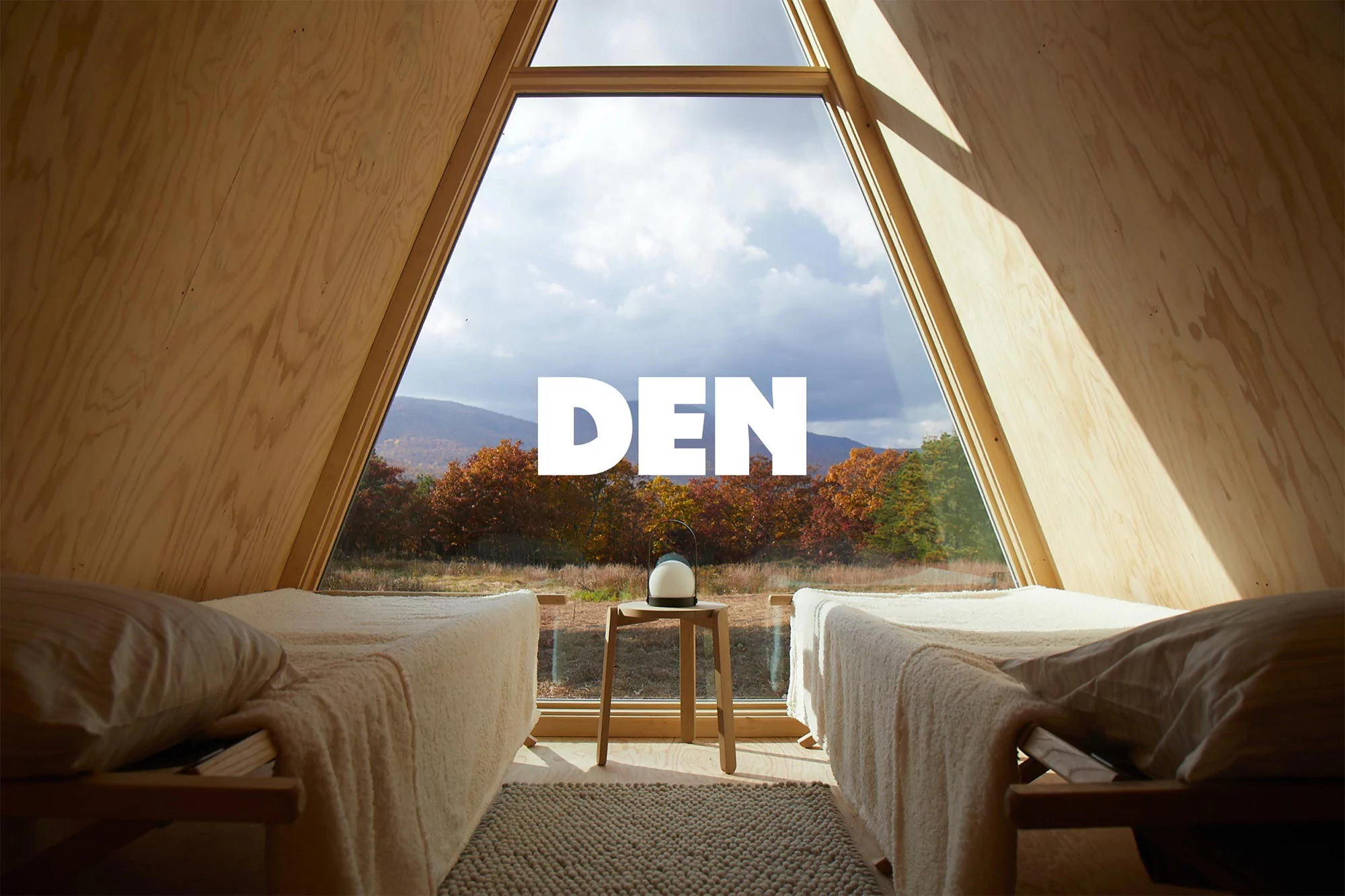A-frame cabin interior with two twin beds facing a large window with a view of trees, mountains, and clouds; minimal decor and natural light.