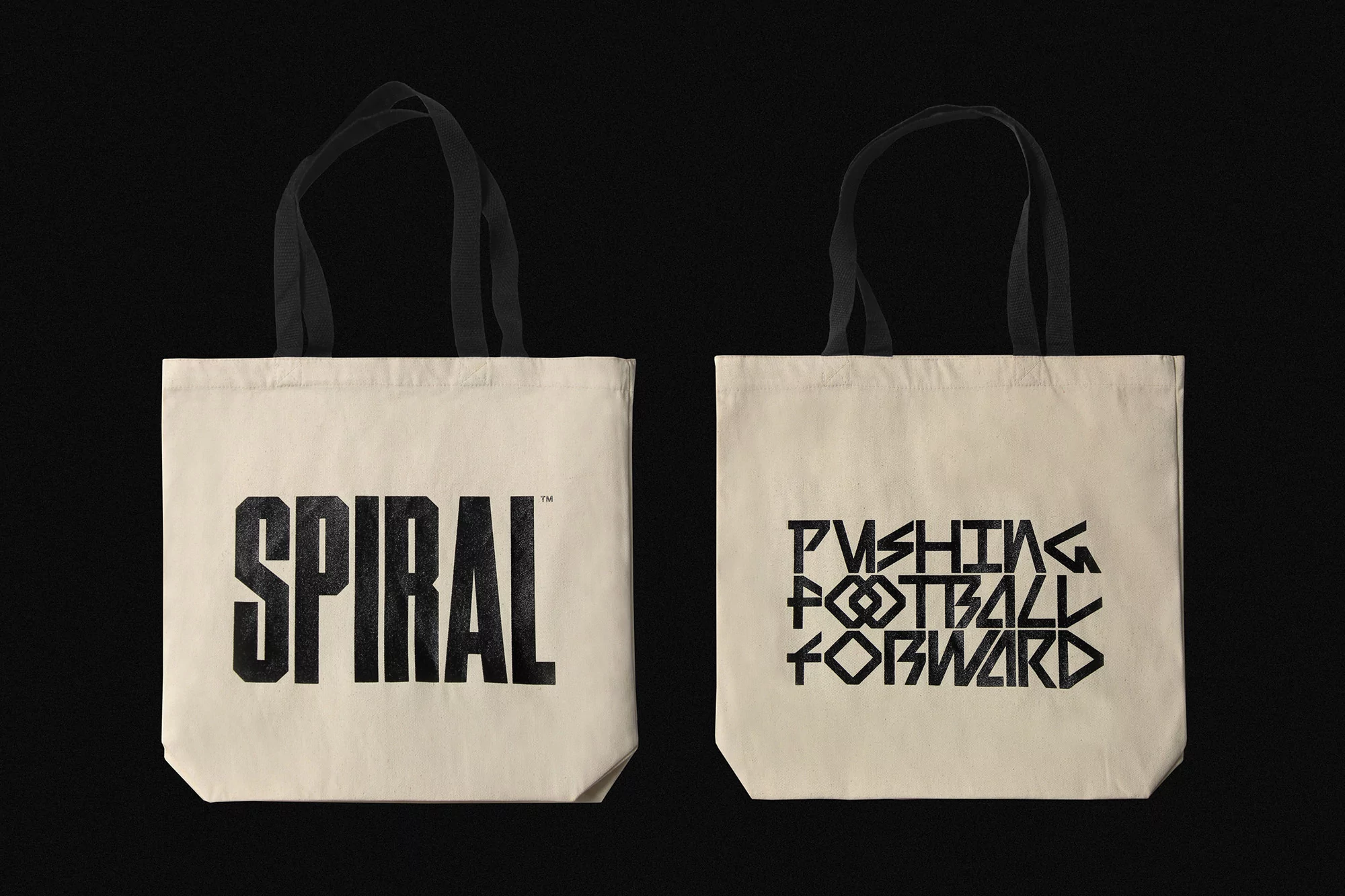 Two cream-colored tote bags, one with the word 'Spiral' in black block letters, the other with 'Pushing Football Forward.'