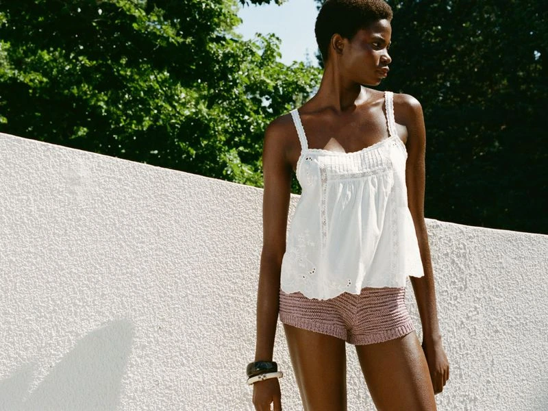 A dark-skinned woman in a white eyelet tank top and mauve shorts stands in an outdoor setting with greenery overhead.