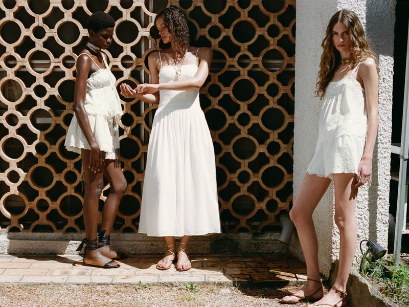Three women stand outdoors on a sunny day, wearing casual white summer dresses and sandals in front of a patterned wall.