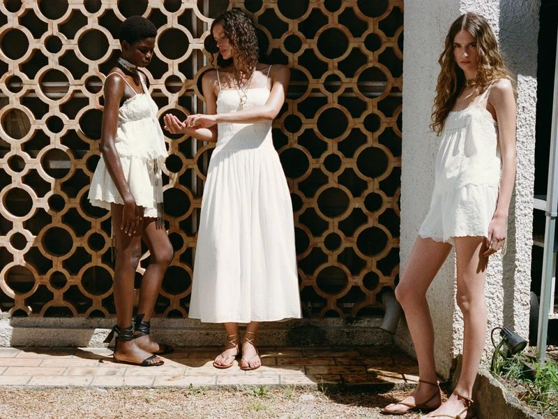 Three women stand outdoors on a sunny day, wearing casual white summer dresses and sandals in front of a patterned wall.