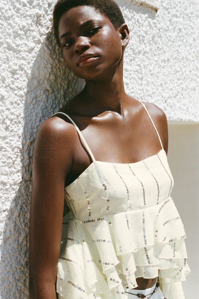 Dark-skinned woman poses against a wall in a tiered, ruffle-sleeved blouse with sequin details in an outdoor setting, shot in natural light.