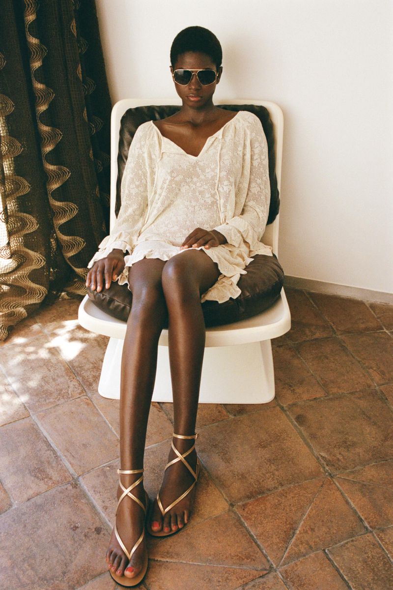Dark-skinned woman in a white dress and sunglasses sits on a chair with tile floors and a curtain visible in the background.