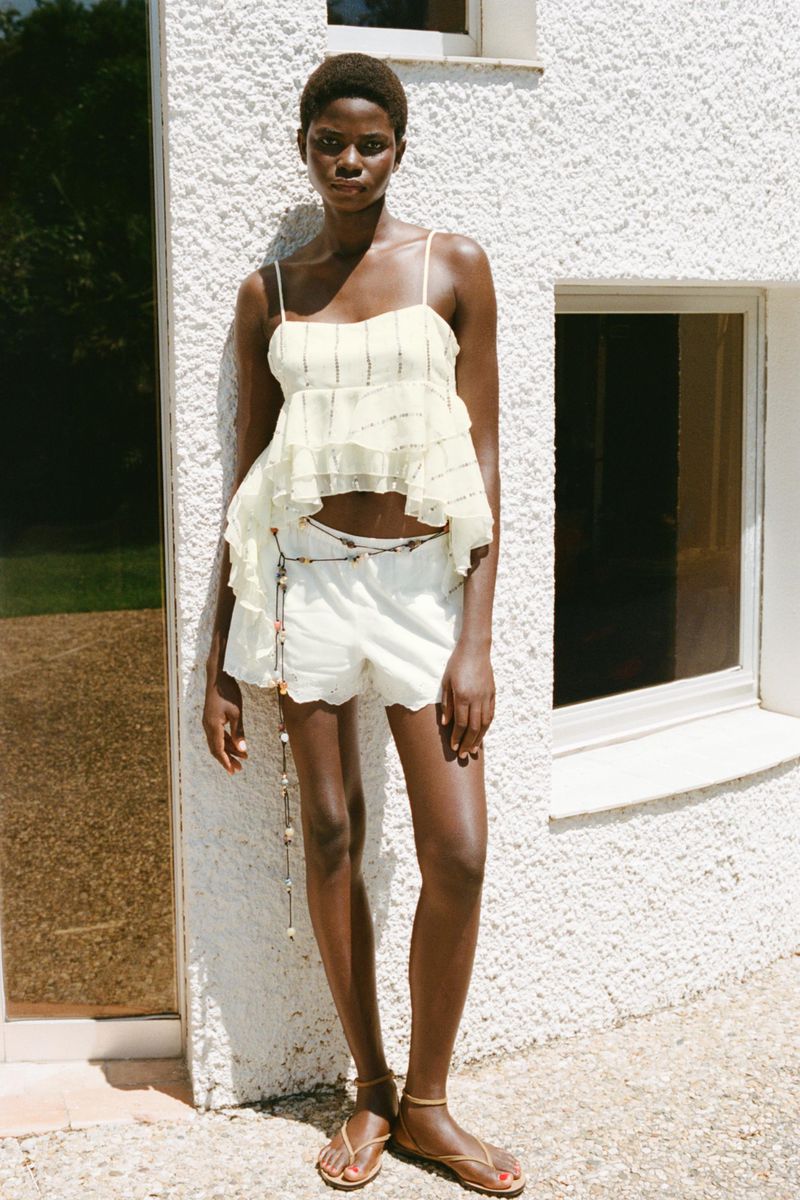 A slender African-American woman poses in front of a white-textured wall, modeling a tiered yellow top and white shorts with strappy sandals.