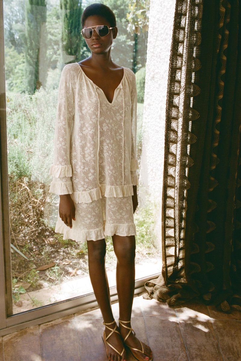 A woman with dark skin stands in front of a window wearing a white floral dress and sunglasses, with a lace curtain to the side.
