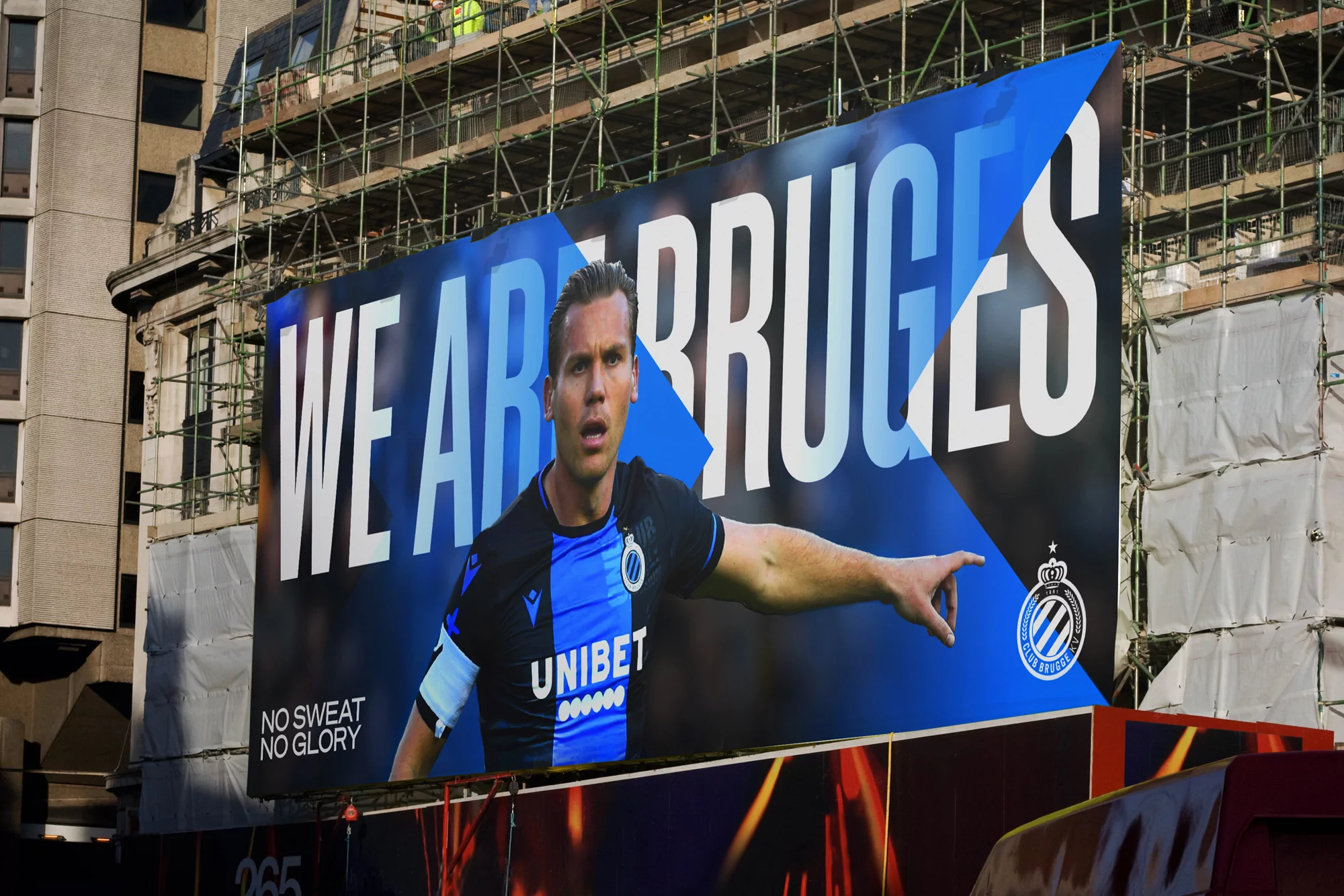 Advertising billboard for Club Brugge soccer team featuring a male athlete in a blue and black uniform in an urban setting.