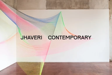 Colorful mesh net installation suspended across a white wall inside a gallery space with exposed concrete ceiling and brown floor.