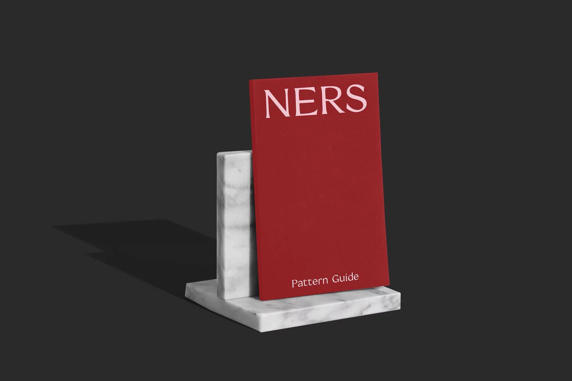 Still life shot of a red 'NERS Pattern Guide' book displayed on a white marble block against a dark grey backdrop.