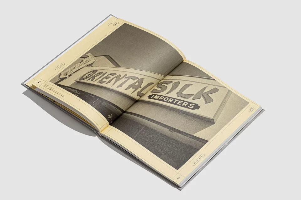 Open book displays a black-and-white photograph of the sign 'Oriental Silk Importers,' presented on a page with muted yellow tones.