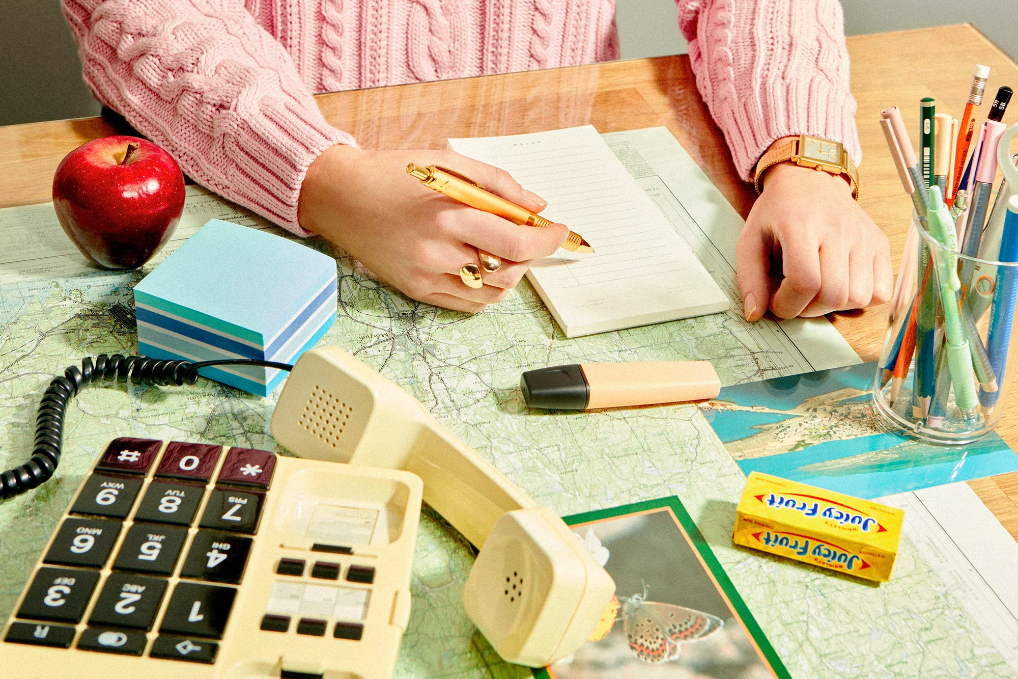 Overhead shot of a person writing on a notepad at a wooden desk covered with a map, next to a vintage phone and Juicy Fruit gum.