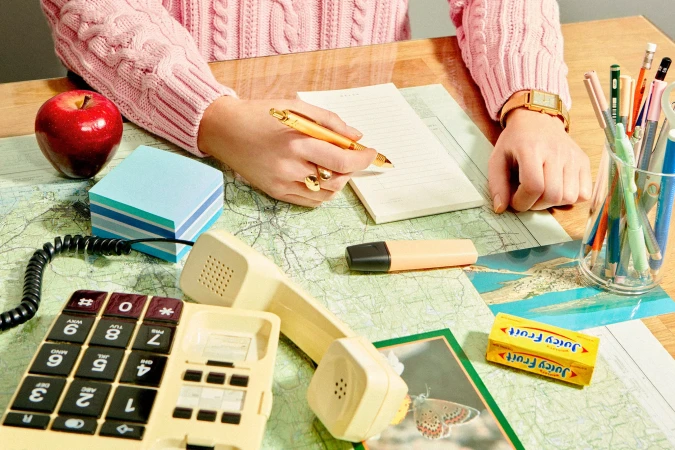 Overhead shot of a person writing on a notepad at a wooden desk covered with a map, next to a vintage phone and Juicy Fruit gum.