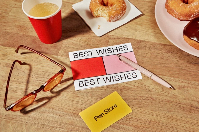 Overhead shot of donuts on a pink plate, a cup of juice, glasses, a pink pen, and a business card on a wooden table.