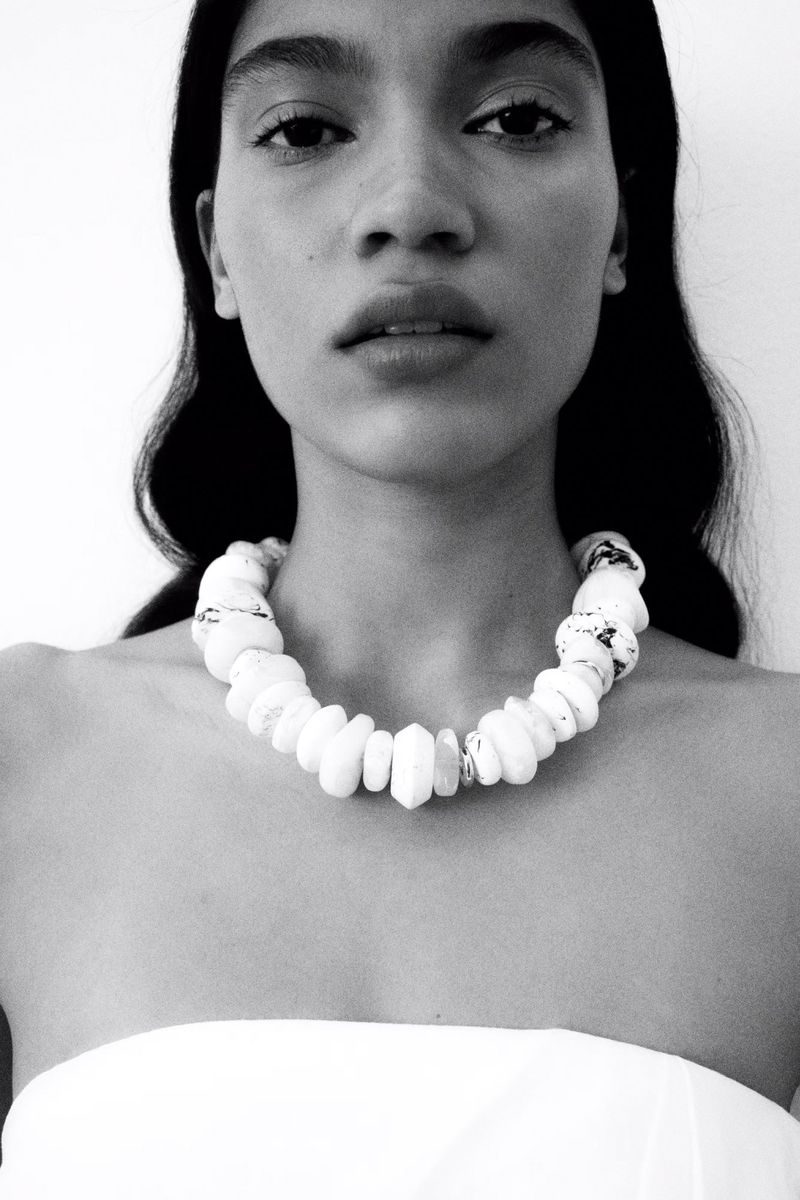 Close-up of a woman wearing a statement necklace with an off-the-shoulder white top against a white background in black and white.