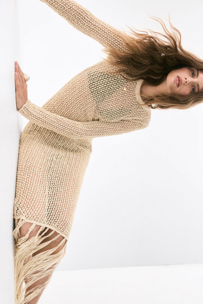 Blonde woman in knit dress with fringe hem posing in a fashion shot against a white wall, looking at the camera with neutral expression.