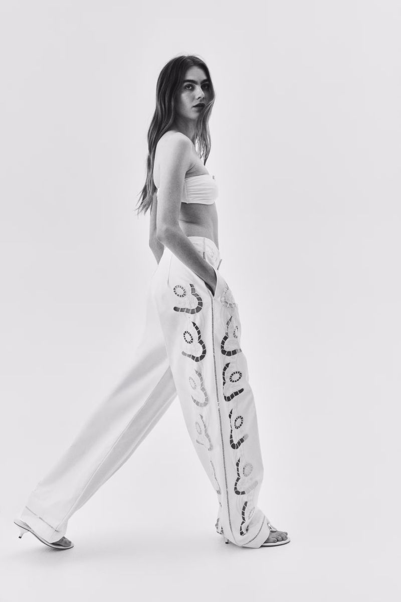 Monochrome studio shot of a young adult woman walking in white top and pants with decorative detail, silver heels, and soft, diffused lighting.