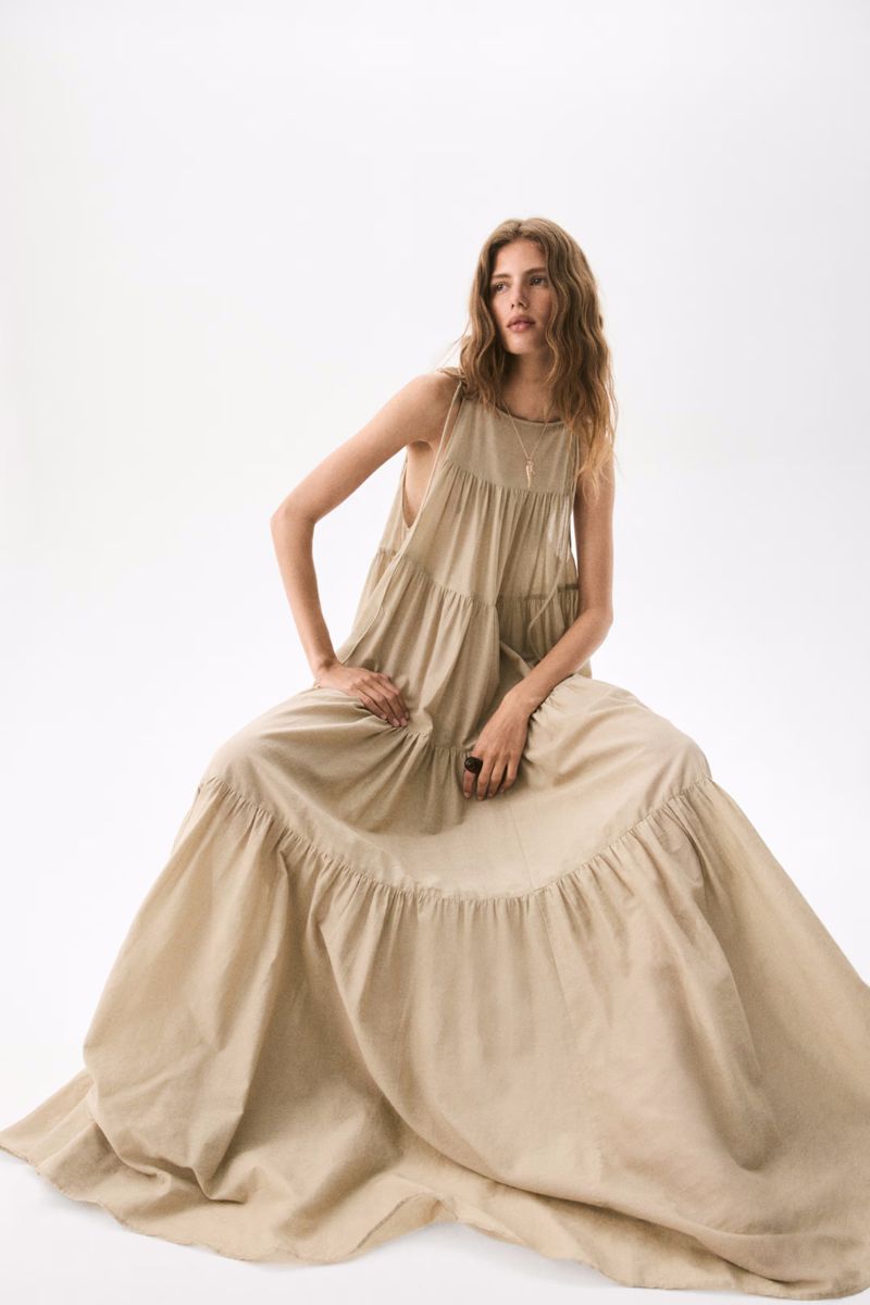 A fashionable adult woman wearing a long, flowing tan dress is seated in a brightly lit photo studio, creating a minimalist fashion portrait.