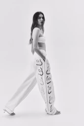 Monochrome fashion editorial image of a female model wearing patterned white trousers and a bandeau top against a plain white studio background.