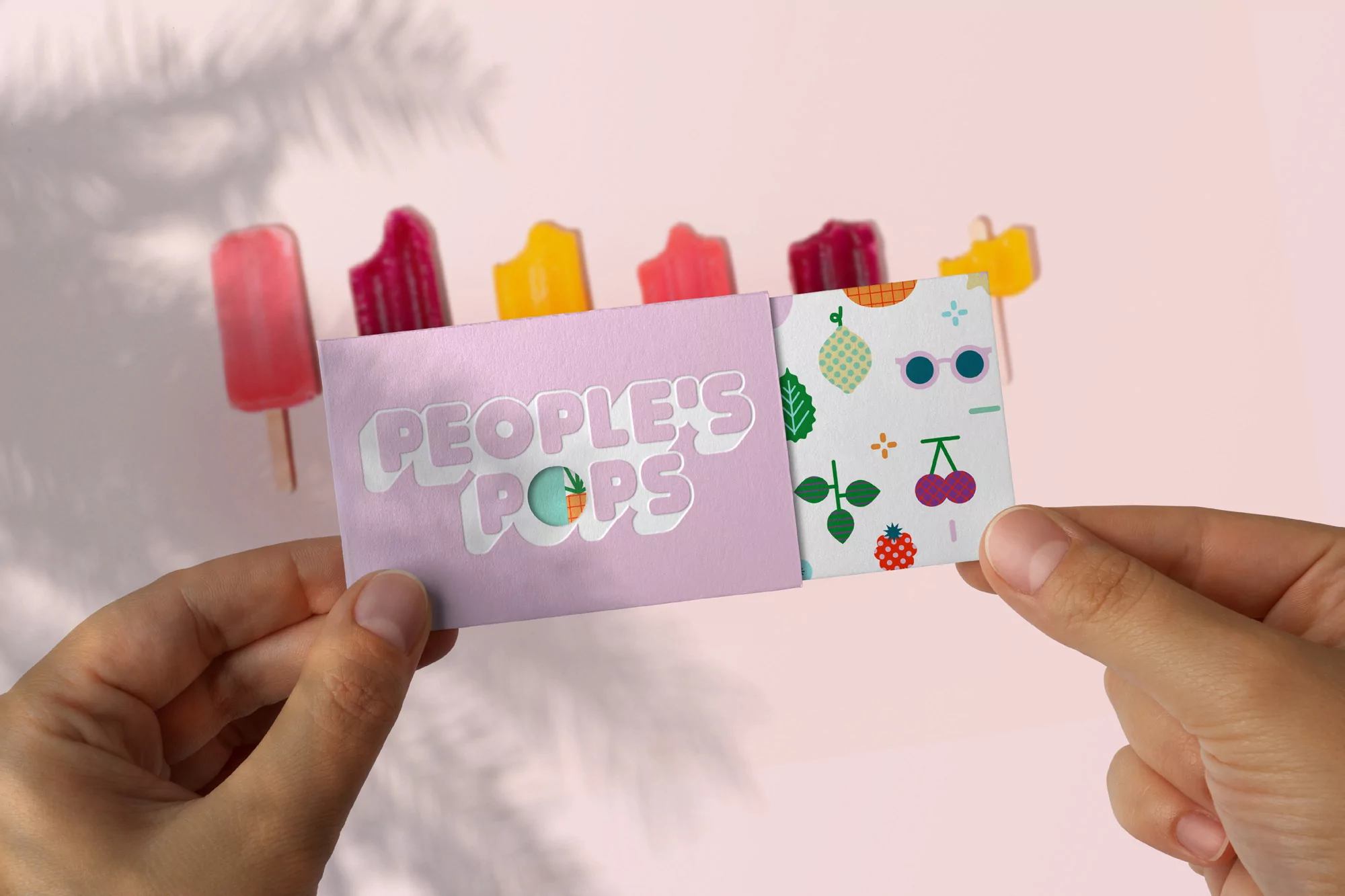 Hands hold People's Pops business card with popsicles in background against pale pink surface with leaf shadows, studio lit scene.