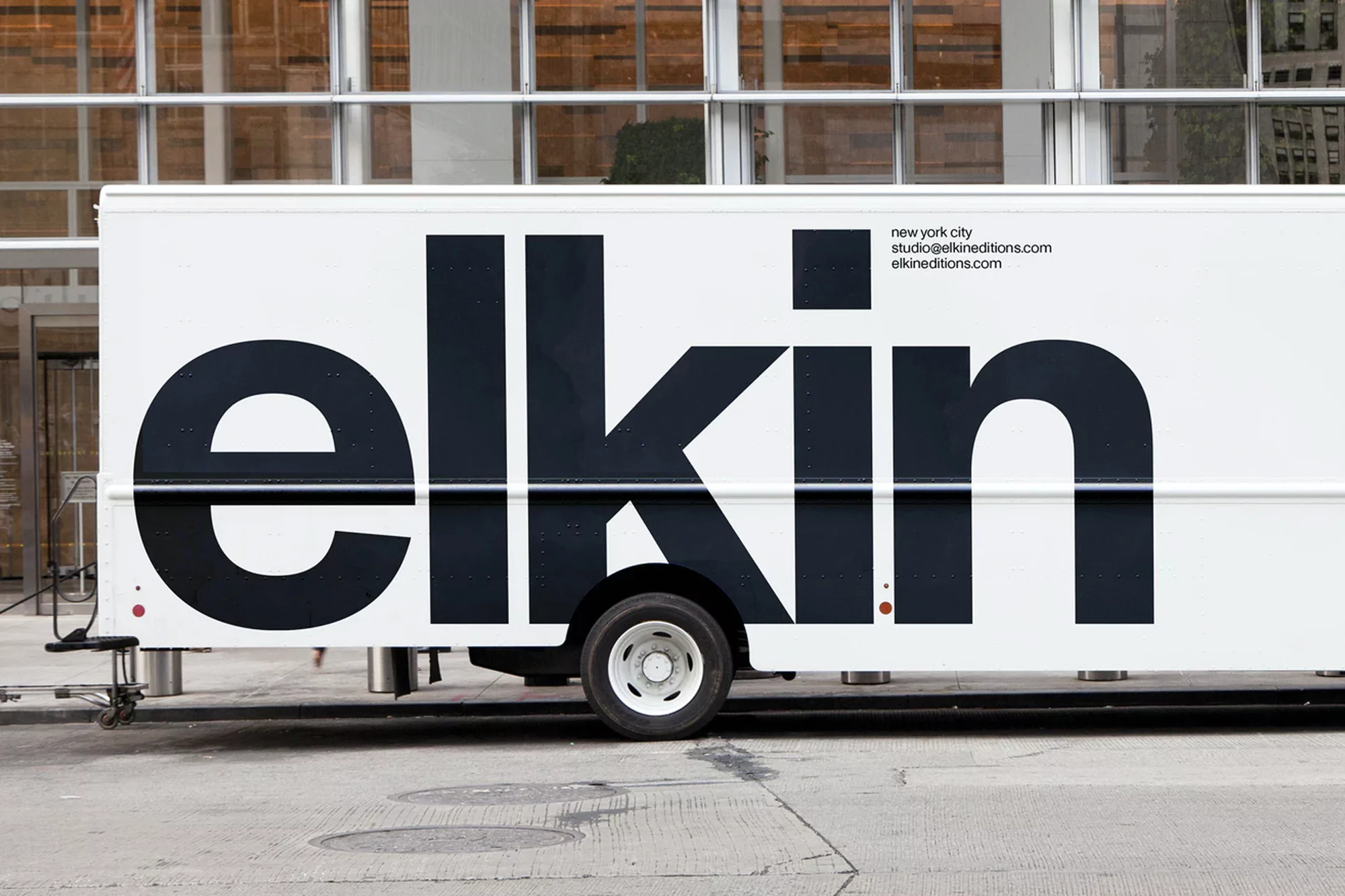 A white box truck with the brand name 'elkin' in large, bold, black letters parked on a city street in front of a modern building.
