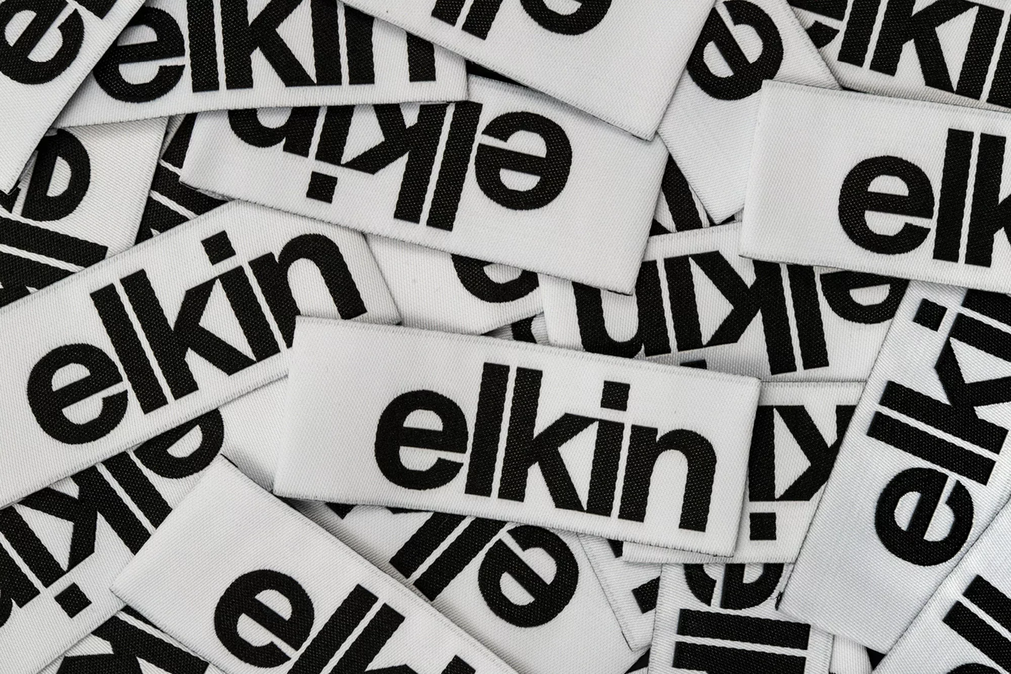 Overhead shot of many white clothing labels with the brand name 'elkin' in black text scattered across the frame.