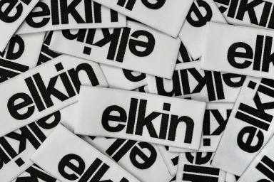 A flat lay pile of woven white clothing labels featuring the bold black Elkin brand logo.