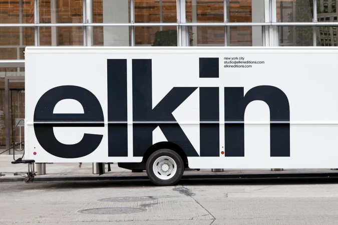 Side view of a white delivery truck featuring large bold dark typography spelling elkin and contact details parked on a city street.