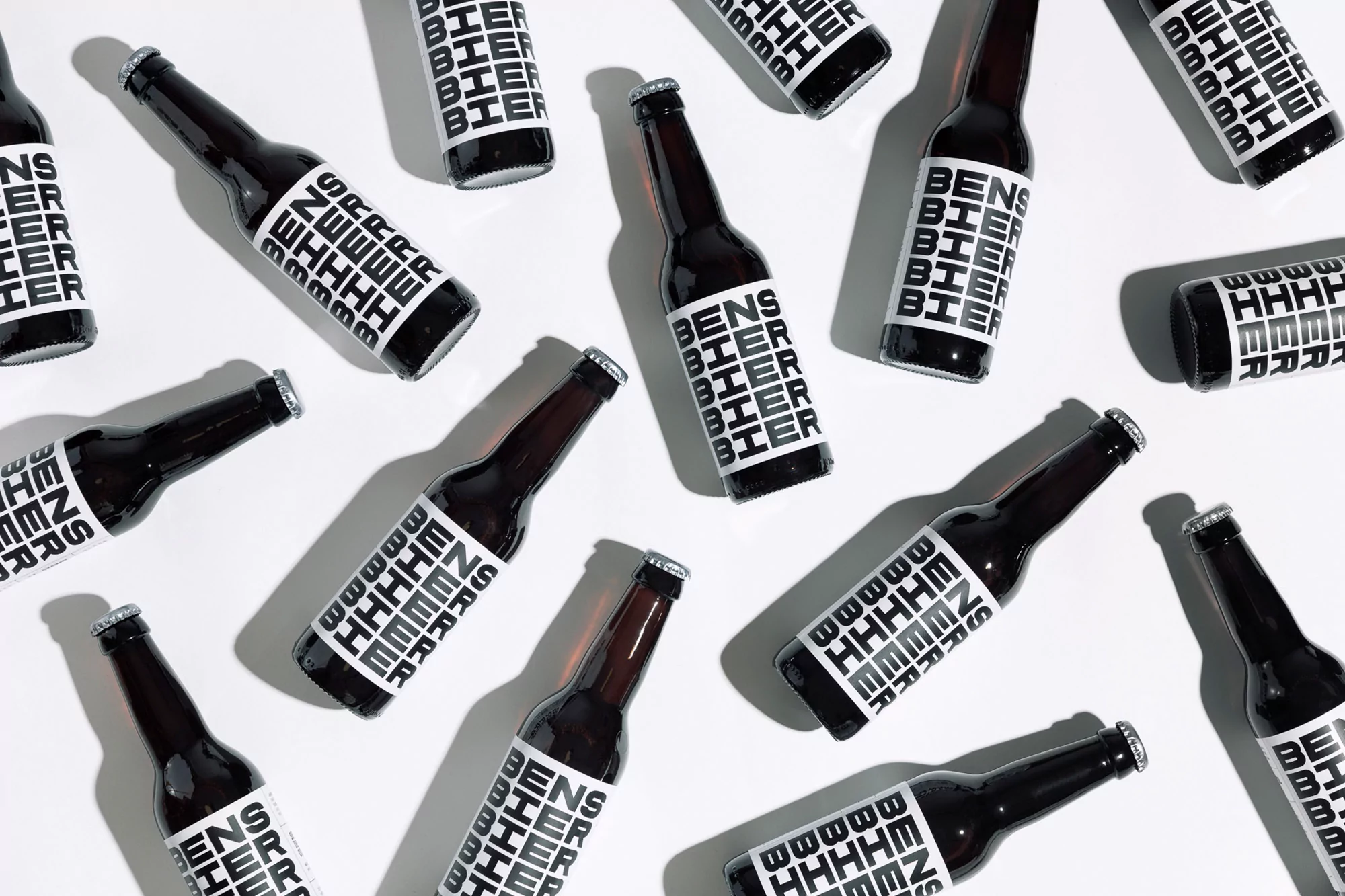Multiple dark beer bottles with white labels angled on a white surface, creating a pattern of commercial drinks in a still life.