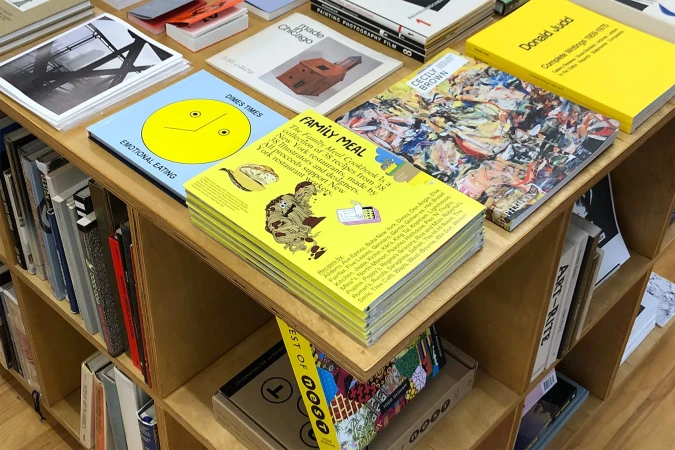 Display of art and design books on a wooden shelf, featuring titles like 'Family Meal,' 'Emotional Eating,' and 'Donald Judd.'